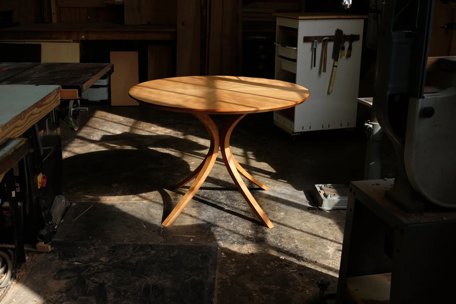 A round wooden table with three curved legs in a workshop, illuminated by sunlight and shadows